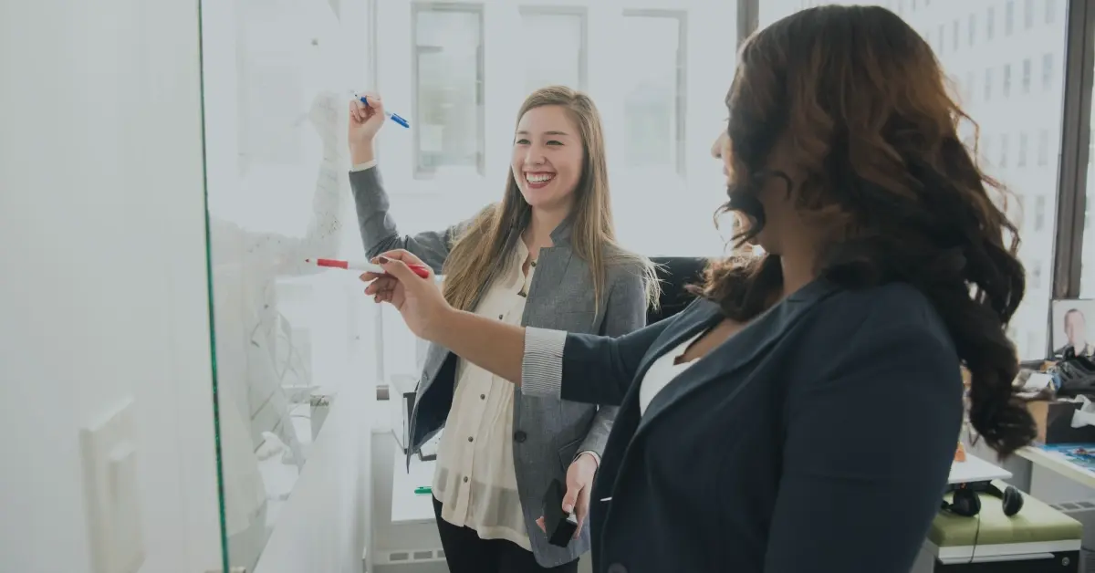 Two executive women claim adjusters learning and writing on a whiteboard.