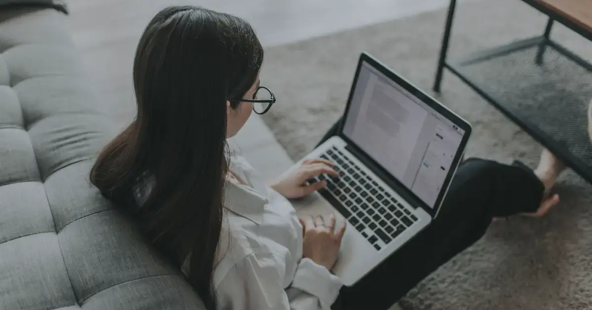 woman with dark hair and glasses wearing a white shirt sits on the floor working on her laptop